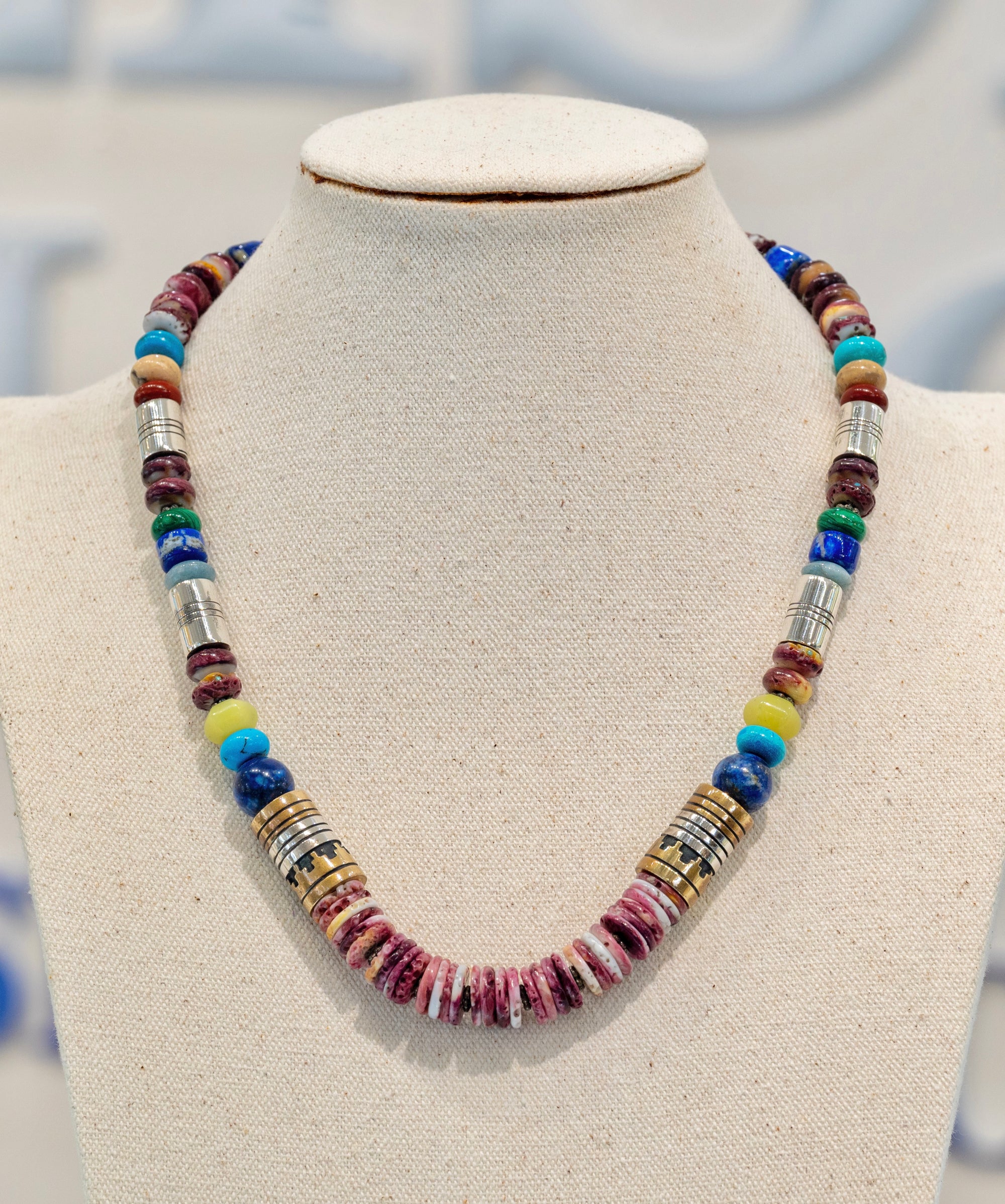 Tommy Singer Navajo Necklace available at Talisman Collection Fine Jewelers in El Dorado Hills, CA and online. Stats: Adorn yourself with a masterpiece of culture and tradition. This necklace features strikingly layered stones, handcrafted by the renowned Navajo artist, Tommy Singer. A vibrant blend of purple spiny oyster shell, lapis, turquoise, and pale serpentine, delicately accented by sterling silver and red brass, perfectly symbolizes the rich cultural heritage of the Southwest.